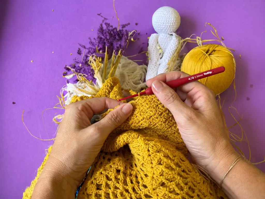 Mains travaillant sur un projet de crochet jaune avec des fils et une poupée à côté sur un fond violet.