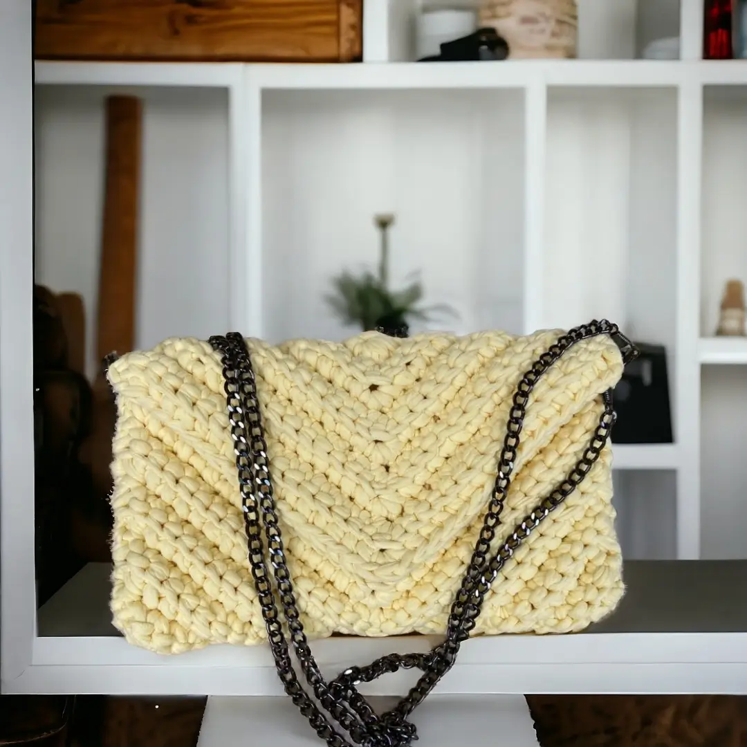 mes-crations-crochellem-galerieweb (1) "Sac à main en crochet de couleur crème avec une bandoulière en chaîne noire, posé sur une table contre un fond d'étagère blanche."