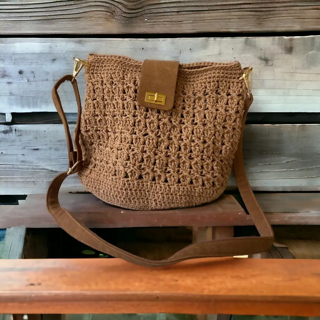 mes-crations-crochellem-galerieweb (10) Sac seau en crochet marron avec fermeture à boucle et bandoulière ajustable, devant un fond en bois.
