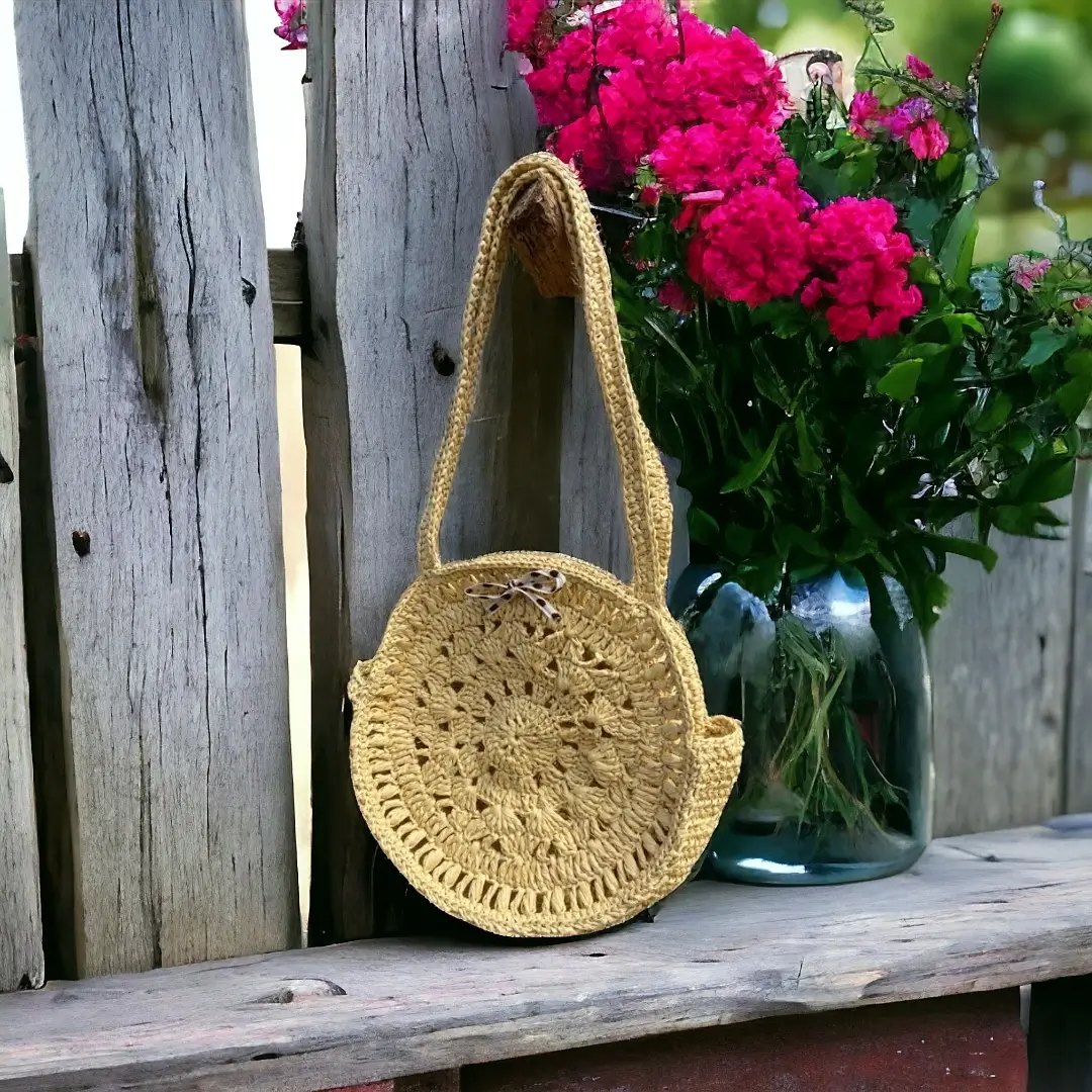mes-crations-crochellem-galerieweb (12) Sac rond en crochet de couleur moutarde suspendu à une clôture en bois, avec des fleurs roses en arrière-plan.