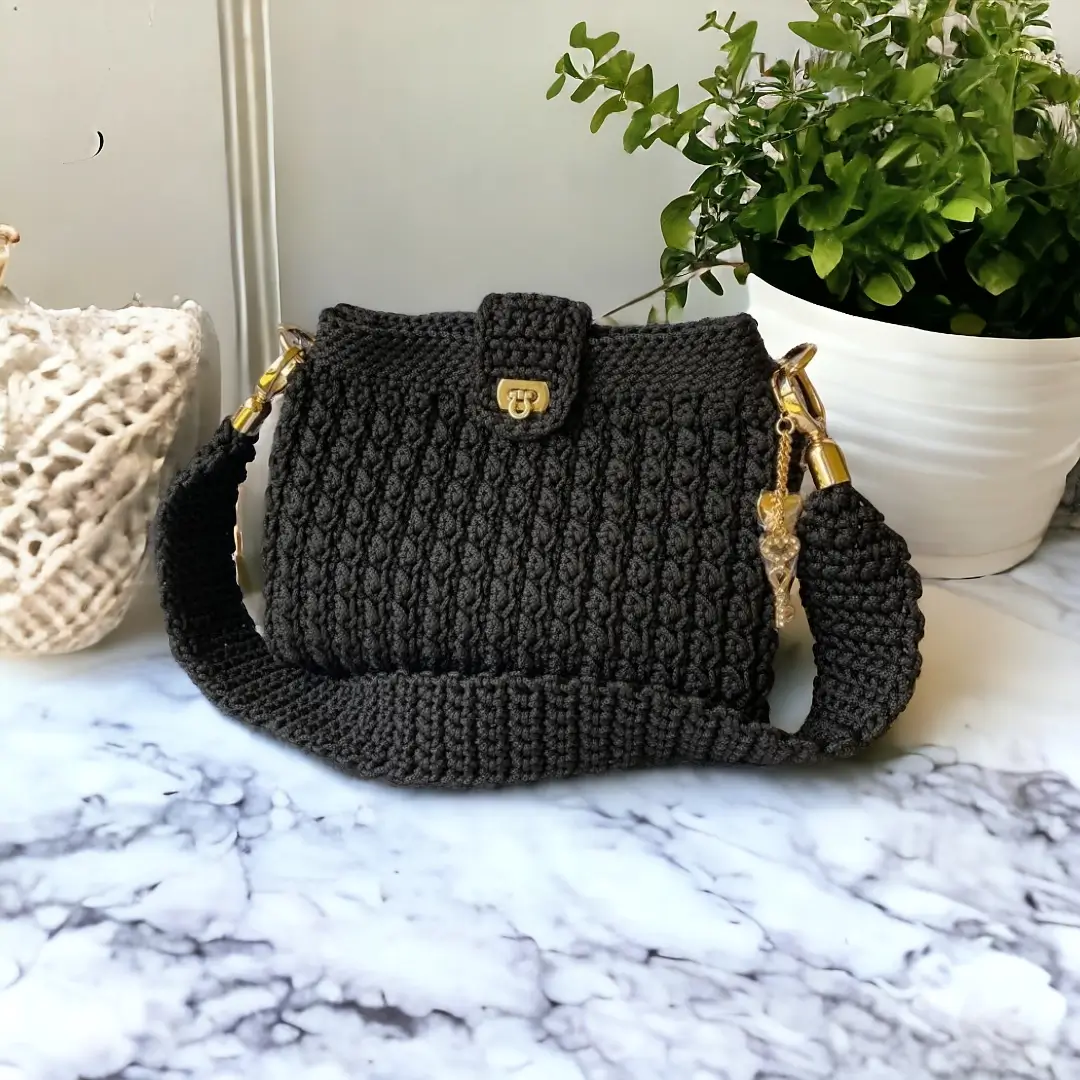 mes-crations-crochellem-galerieweb (15) Sac à main en crochet noir avec détails dorés et breloque en forme de cœur, posé sur une surface en marbre.