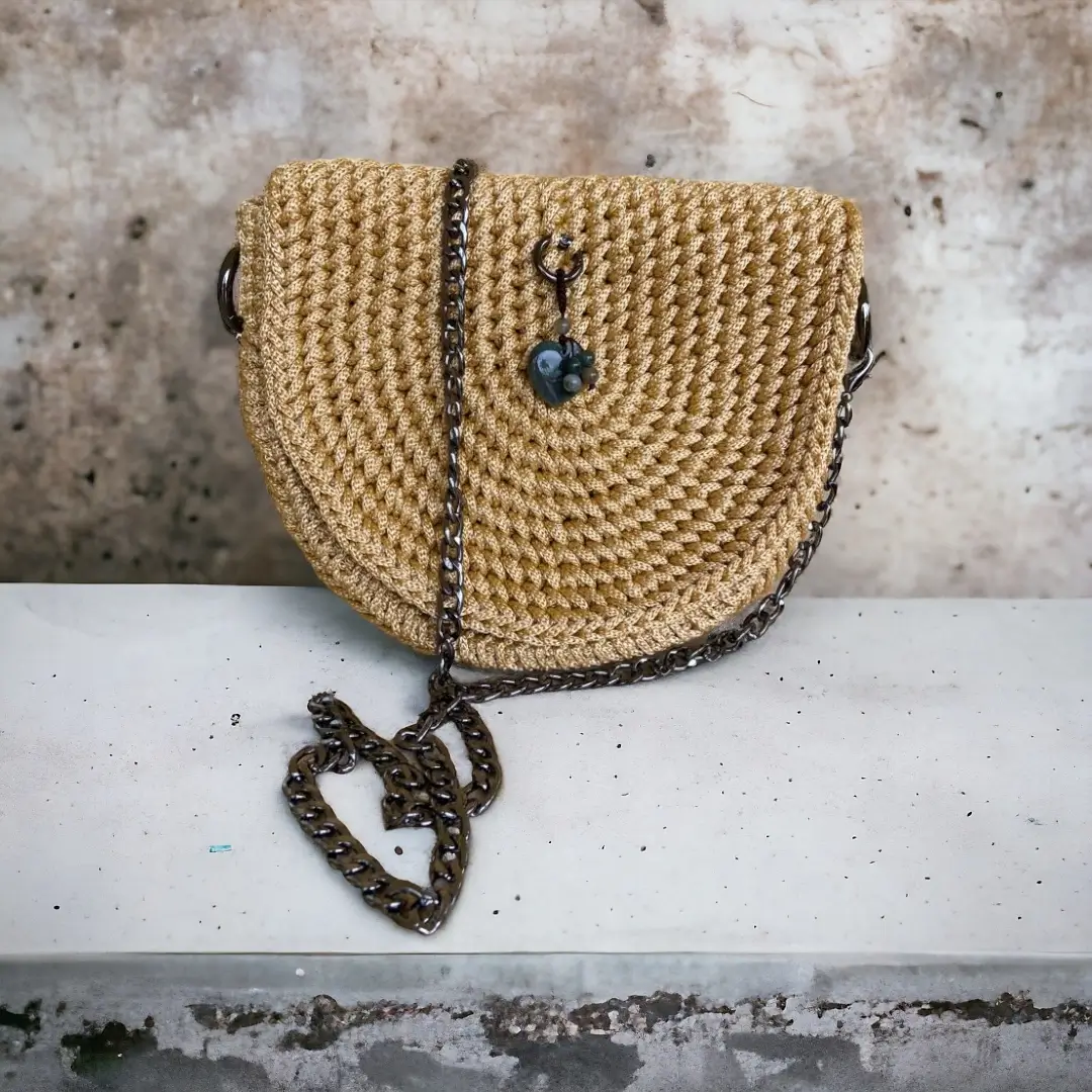mes-crations-crochellem-galerieweb (16) Sac à bandoulière demi-lune en crochet couleur moutarde avec chaîne brun foncé et fermoir à perle bleue, posé sur un rebord de mur texturé.
