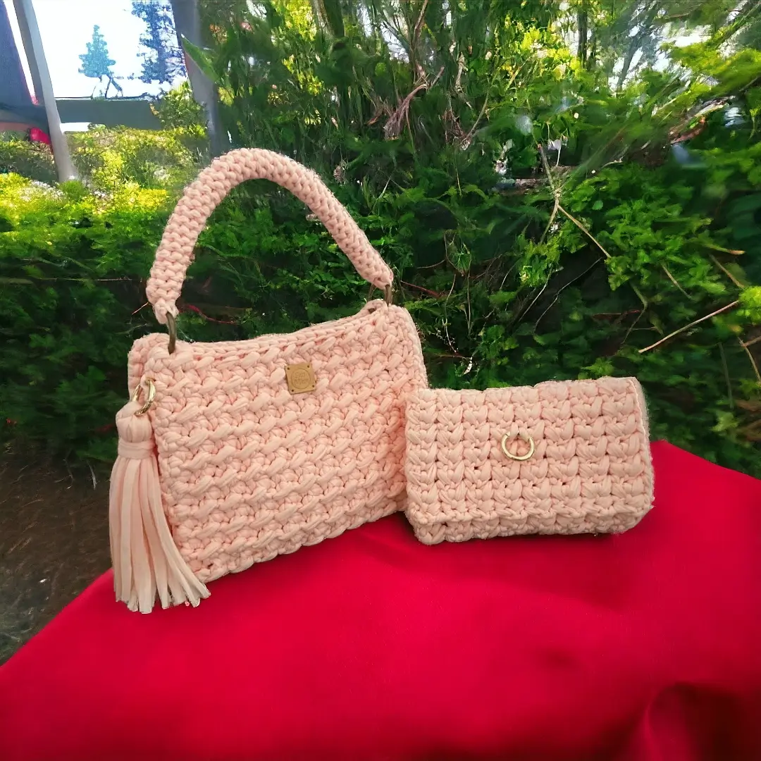 mes-crations-crochellem-galerieweb (17) Ensemble assorti de sac à main et portefeuille en crochet rose pâle avec gland décoratif et fermeture métallique, posé sur un tissu rouge.
