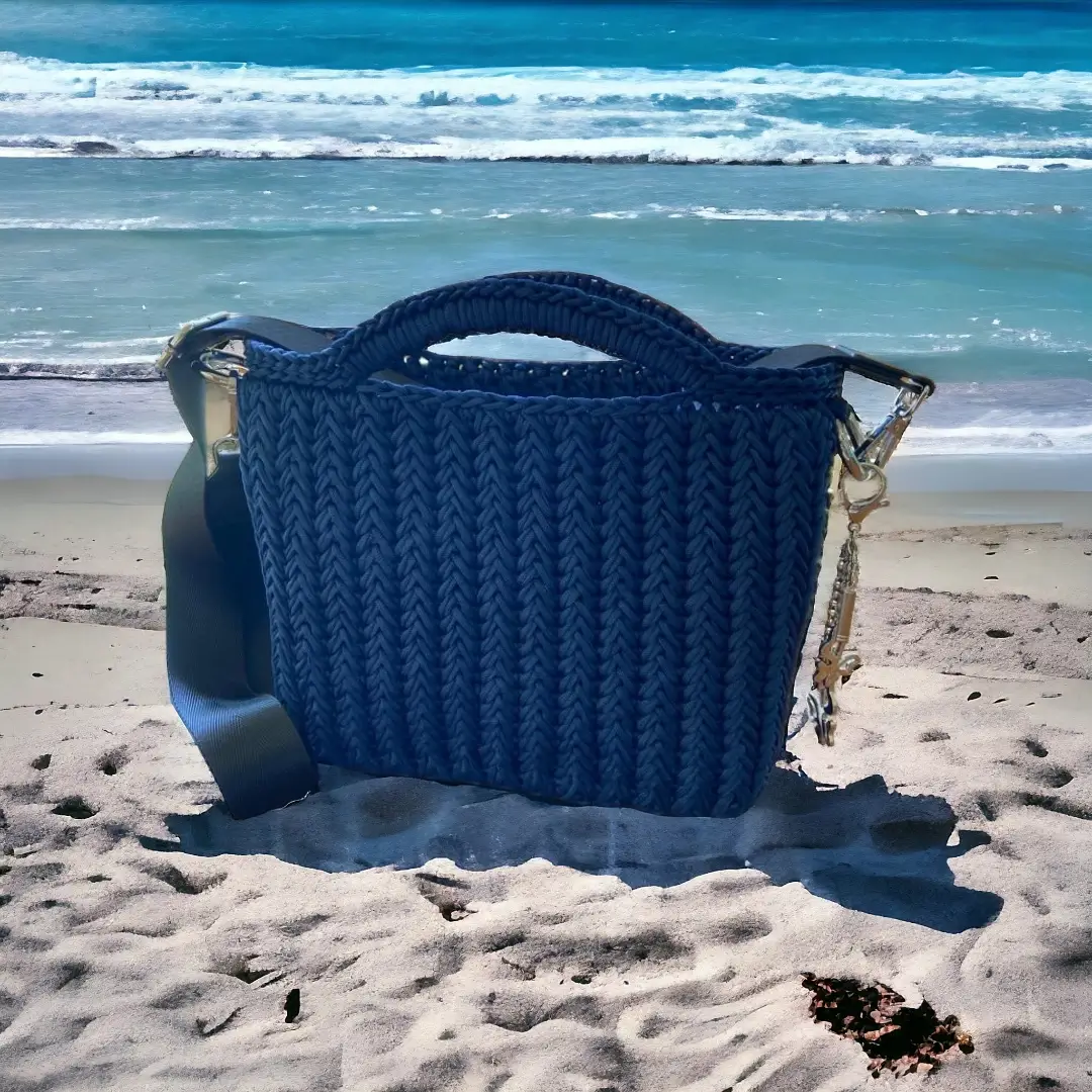 mes-crations-crochellem-galerieweb (3) Sac à main en crochet bleu avec sangle et détails de glands, sur une plage de sable devant la mer.