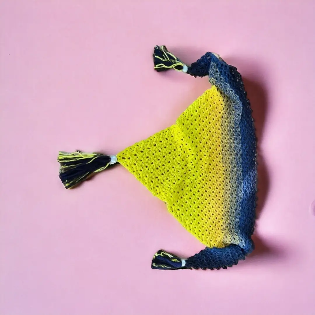 mes-crations-crochellem-galerieweb (6) Châle triangulaire en crochet dégradé du jaune au bleu avec des pompons sur les pointes, présenté sur un fond rose.