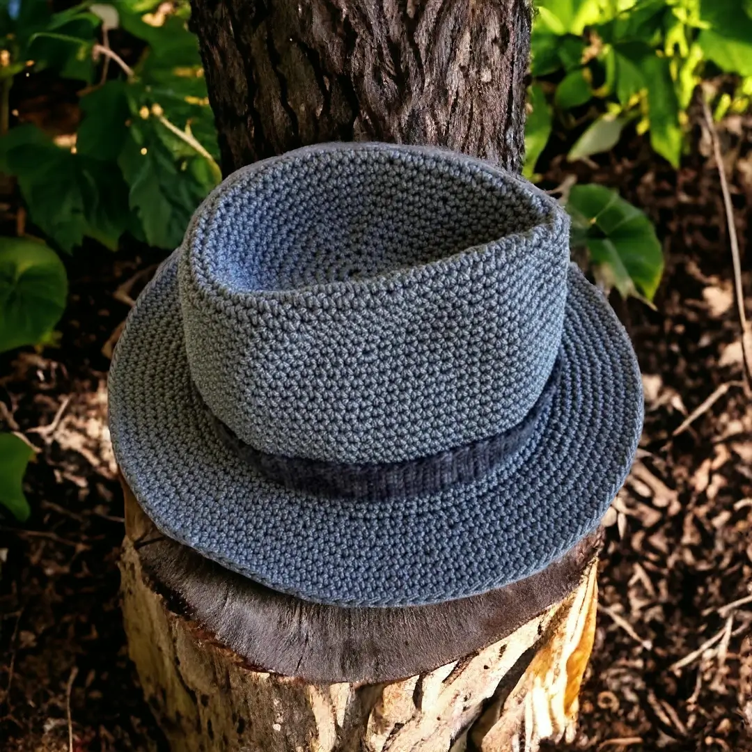 mes-crations-crochellem-galerieweb (8) Chapeau Fedora en crochet gris posé sur un tronc d'arbre avec des feuilles vertes en arrière-plan.