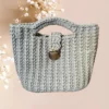 Un sac panier en crochet de couleur gris clair, orné d'une fermeture métallique vintage argenté, combine esthétique traditionnelle et praticité moderne, vue de face